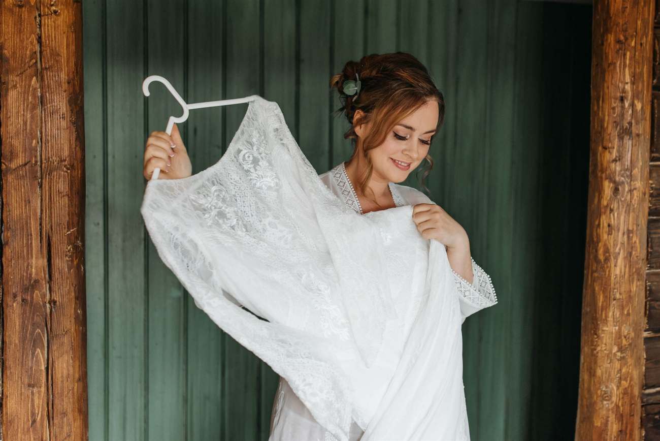 Eine junge Frau präsentiert ein großes, weißes Spitzenkleid vor einem Hintergrund mit vertikalen Holzpfosten.