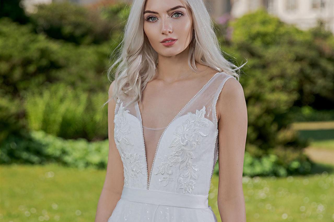 Eine junge Frau in einem weißen, eleganten Kleid steht in einer grünen Parkanlage vor einem historischen Gebäude.
