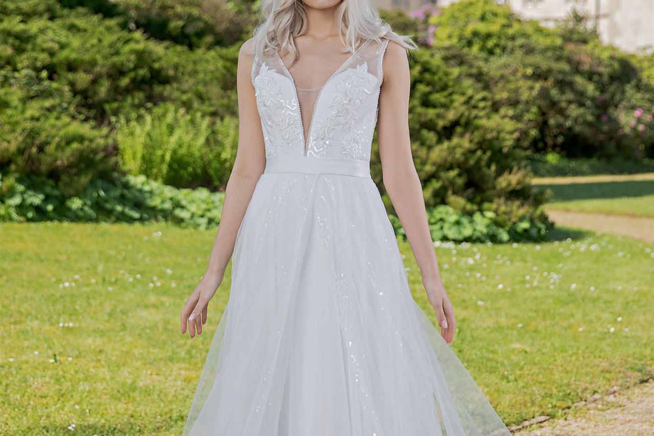 Eine junge Frau in einem fließenden weißen Kleid steht auf einer grünen Wiese vor einem historischen Gebäude.