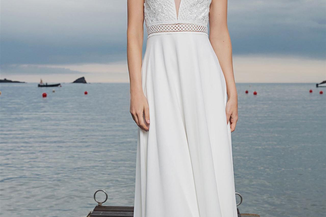 Eine Frau in einem weißen, eleganten Kleid steht auf einem Holzpier vor einem weiten Meer.