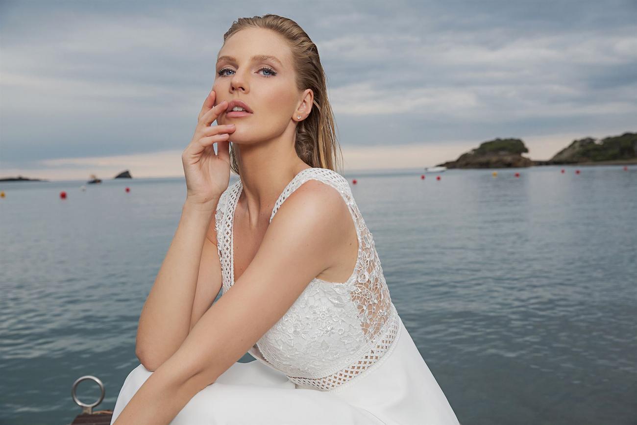 Eine Frau in einem weißen Spitzenkleid posiert elegant am Ufer vor einer weiten Wasserfläche mit nebelverhangener Küste im Hintergrund.