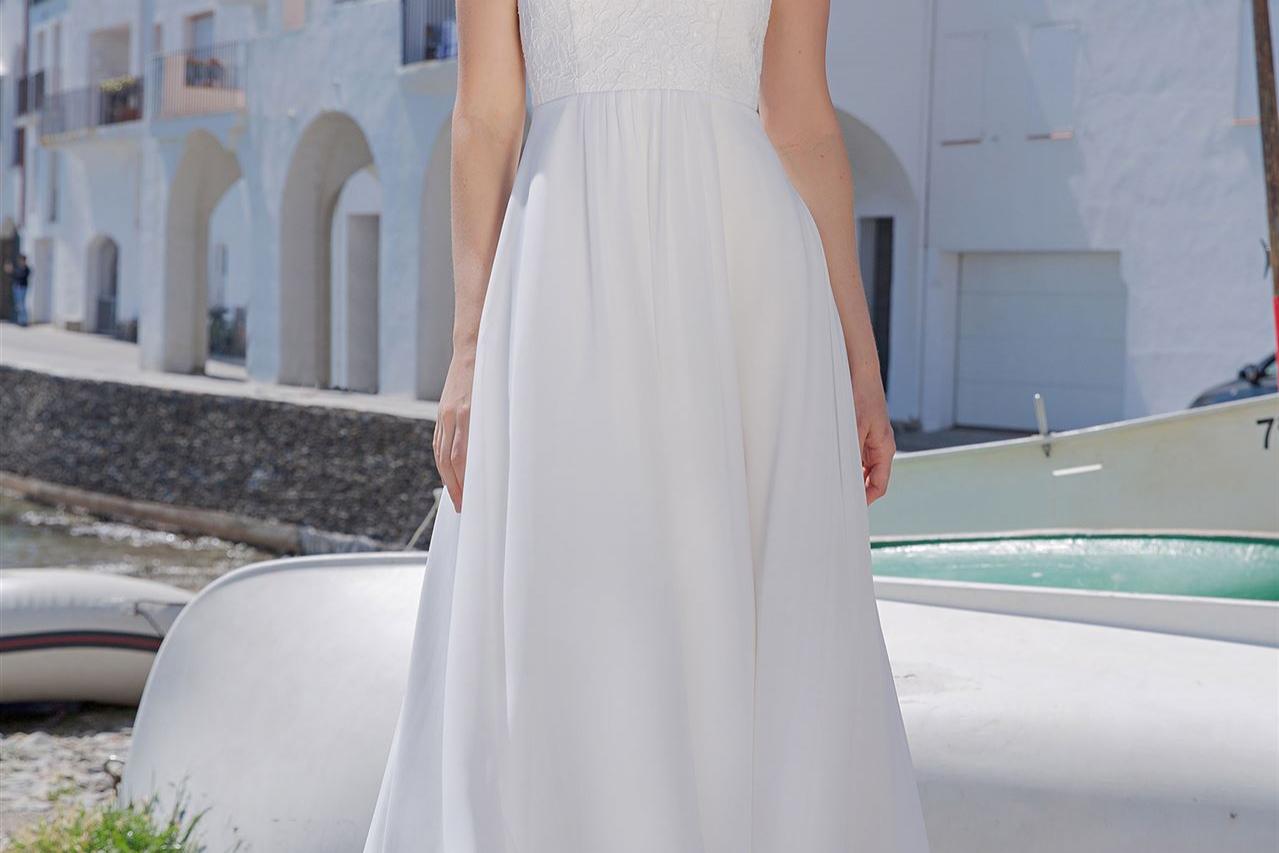 Eine Frau in einem weißen, fließenden Kleid steht auf einem hölzernen Steg vor einer mediterran anmutenden Küstenbebauung.
