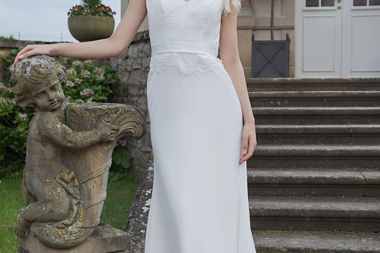 Eine blonde Frau in einem weißen Hochzeitskleid lehnt an einer antiken Skulptur vor einer steinernen Treppe.