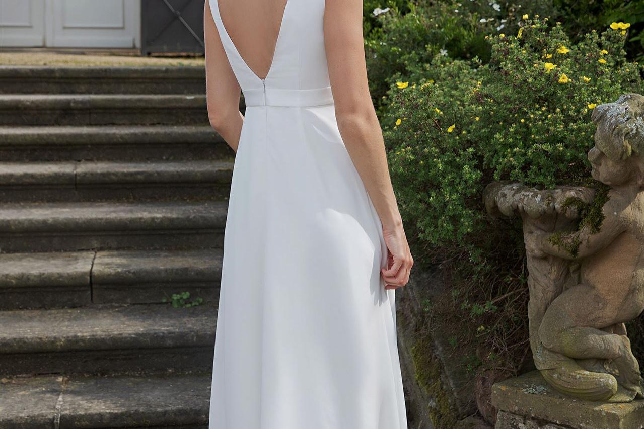 Eine junge Frau in einem weißen, eleganten Kleid mit tiefem V-Ausschnitt steht vor einer historischen steinernen Treppe und üppiger Pflanzenwelt.