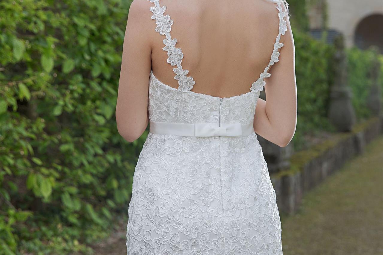 Eine blonde Frau in einem weißen Spitzenkleid steht in einem gepflegten, grünen Garten.