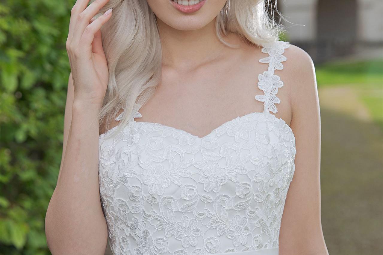 Blonde Frau in einem weißen Spitzenkleid, draußen vor einem Gebäude posierend.