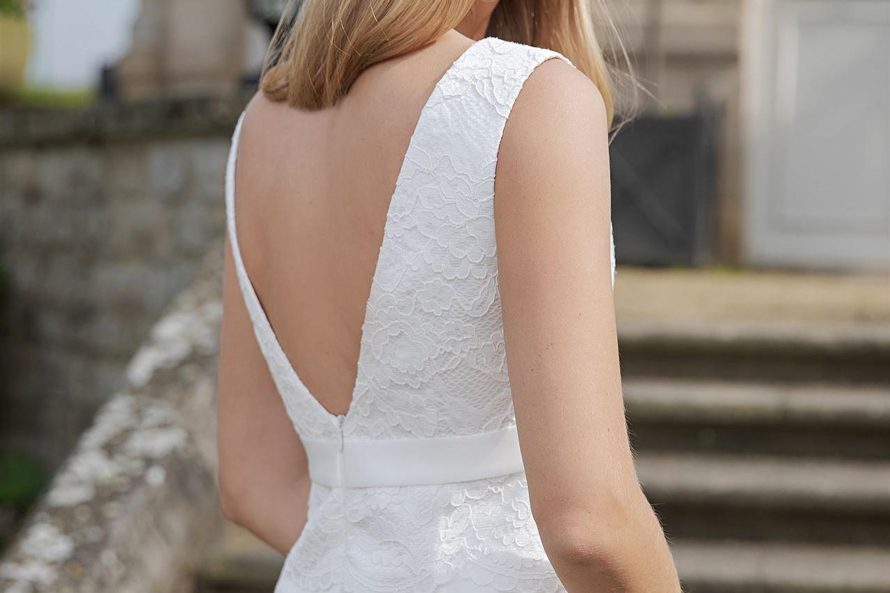 Eine blonde Frau in einem weißen Hochzeitskleid blickt über ihre Schulter auf den Betrachter, während sie vor einer steinernen Treppe steht.