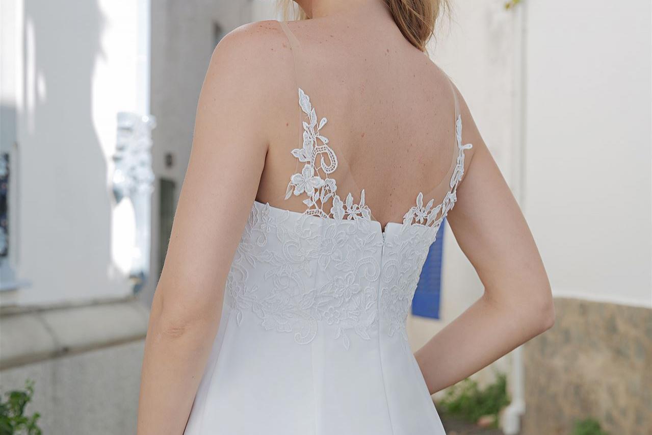 Eine Braut in einem weißen Hochzeitskleid blickt im Freien nach links, umrahmt von Architektur und Grün.