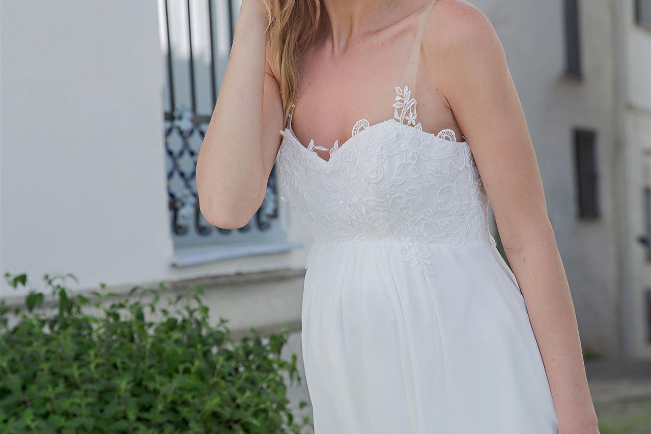 Porträt einer blonden Frau in einem weißen, fließenden Kleid, die vor einem Gebäude mit einem schwarzen Geländer und grünen Pflanzen posiert.
