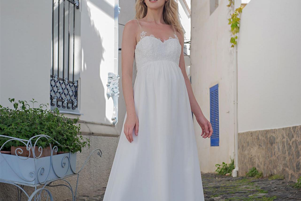 Eine blonde Frau in einem langen, weißen Kleid steht elegant vor einer mediterranen Architektur mit weißen Wänden und blauen Akzenten.
