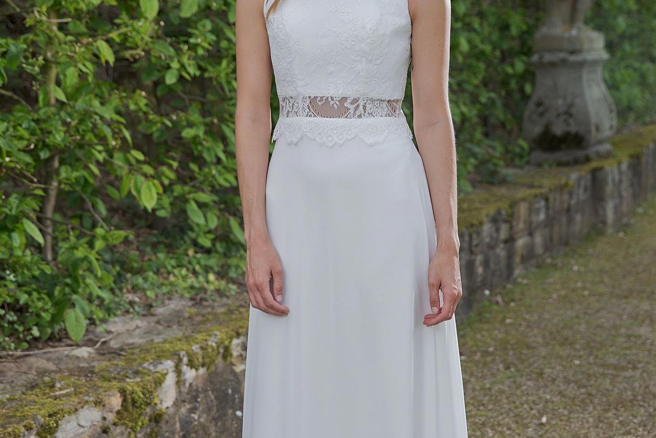 Eine junge, lächelnde Frau in einem langen, weißen Kleid steht vor einem üppär grünen Parkhintergrund.