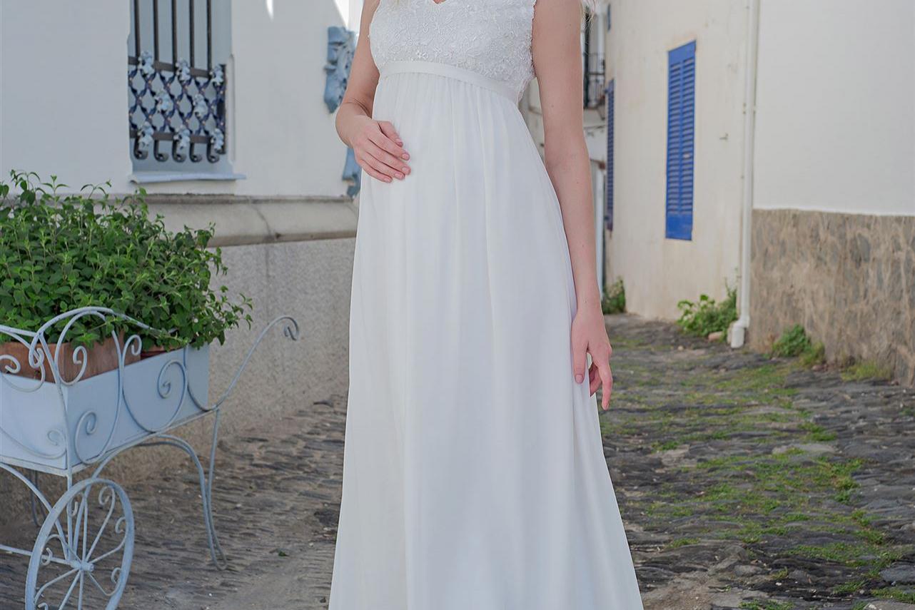 Eine junge Frau in einem fließenden weißen Kleid steht vor einem Hintergrund aus historischen, weiß getünchten Gebäuden.