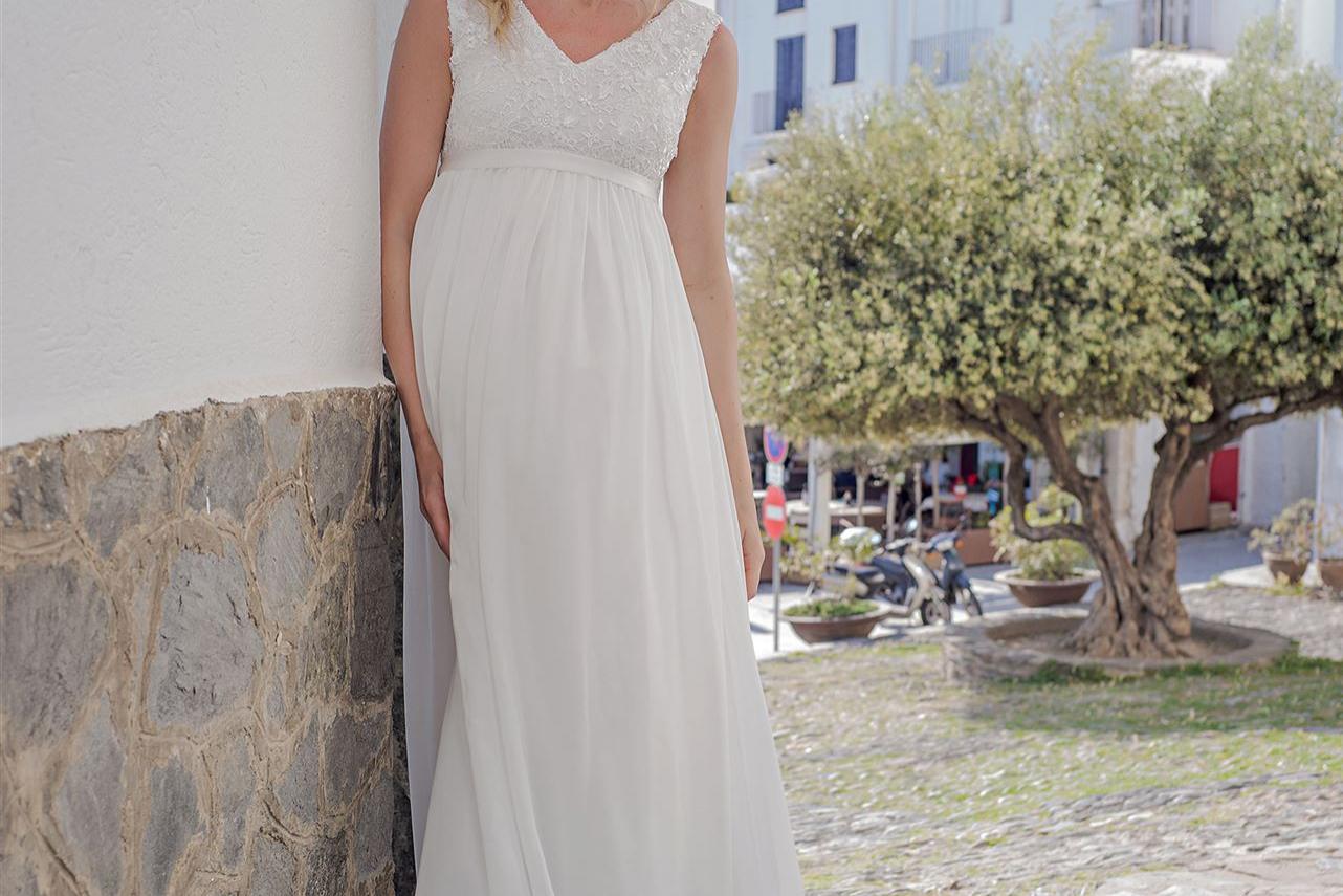 Eine schwangere Frau in einem weißen Kleid lächelt vor einer sonnendurchfluteten Kulisse mit mediterraner Architektur.