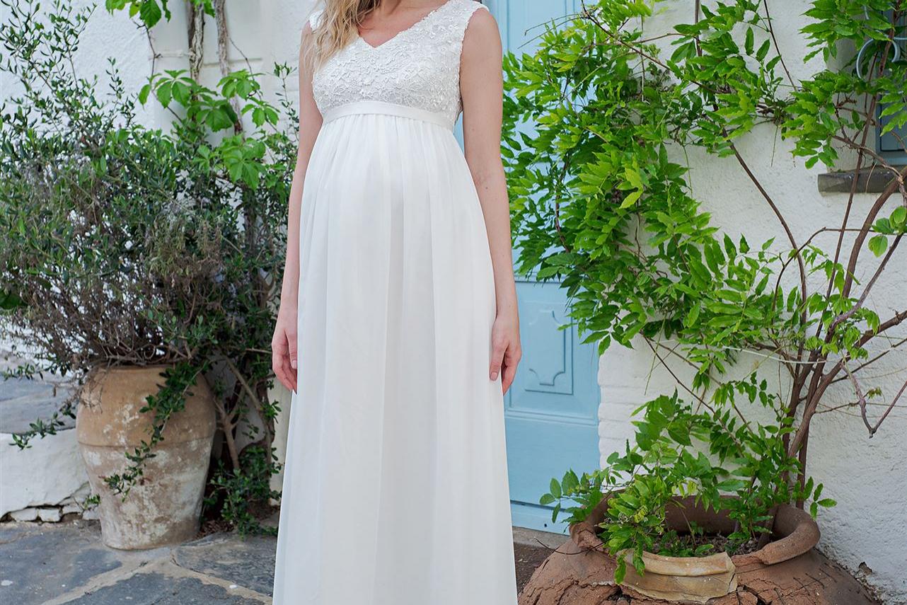 Eine junge Frau in einem langen weißen Kleid steht in einem mediterranen Hof vor einer leuchtend blauen Tür, umgeben von üppigem Grün.