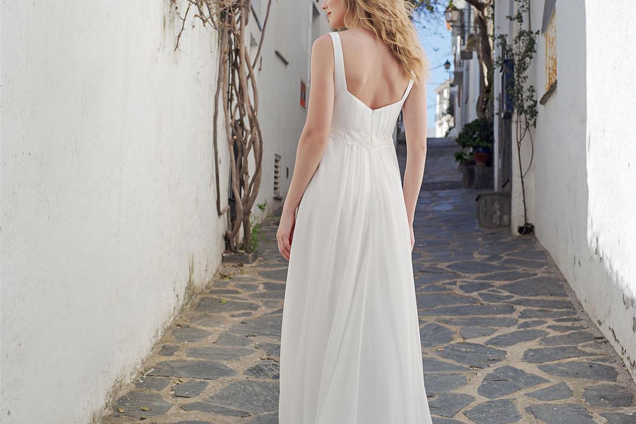 Eine Frau in einem fließenden weißen Sommerkleid geht durch eine enge, von Blumen geschmückte, weißgetünchte Gasse.