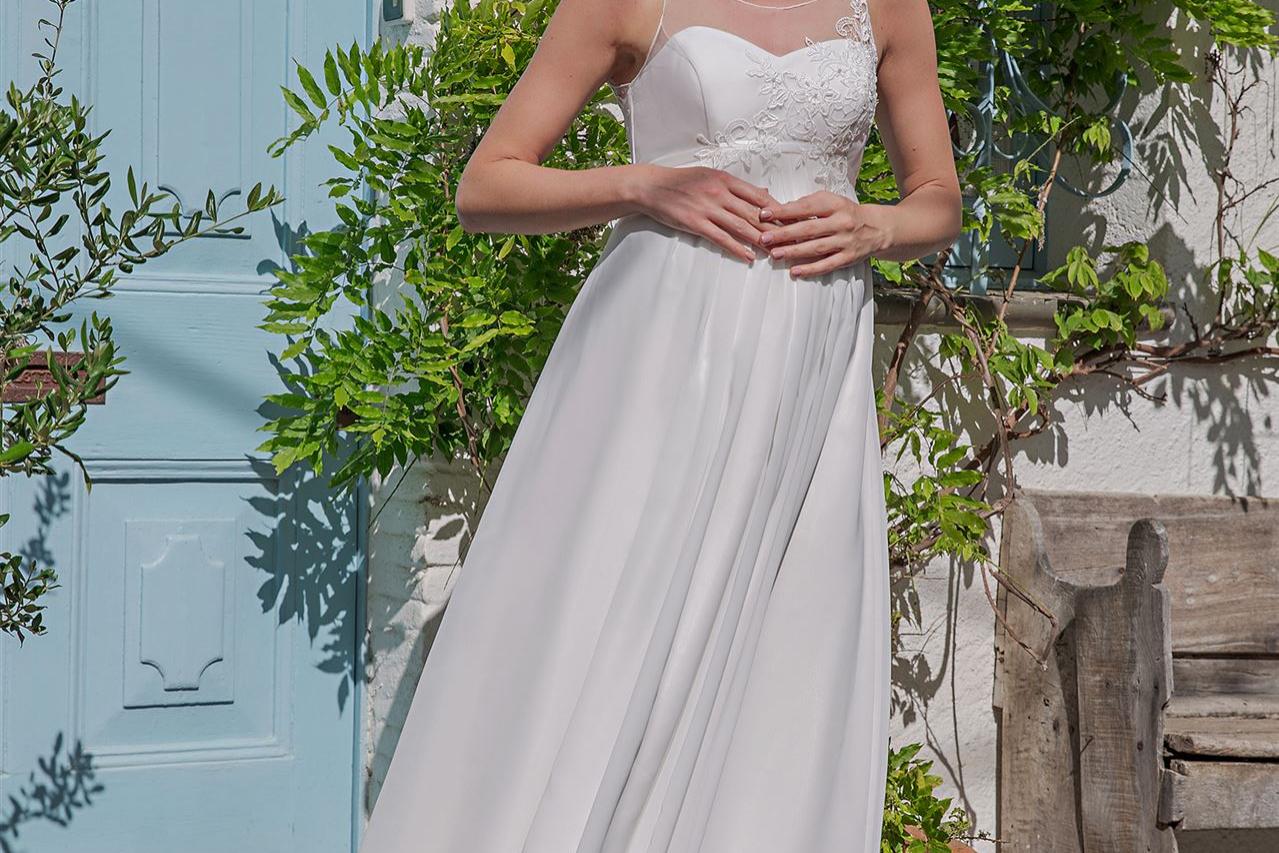 Eine junge Frau in einem weißen Kleid steht draußen zwischen einer hellblauen Tür und dichtem, grünem Laubwerk.