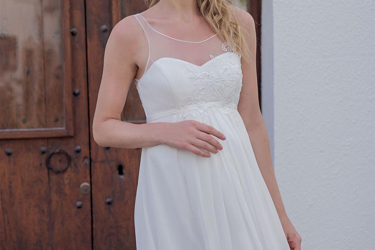 Schwangere Frau in einem weißen Kleid steht lächelnd vor einem Paar dunkler Holztüren.