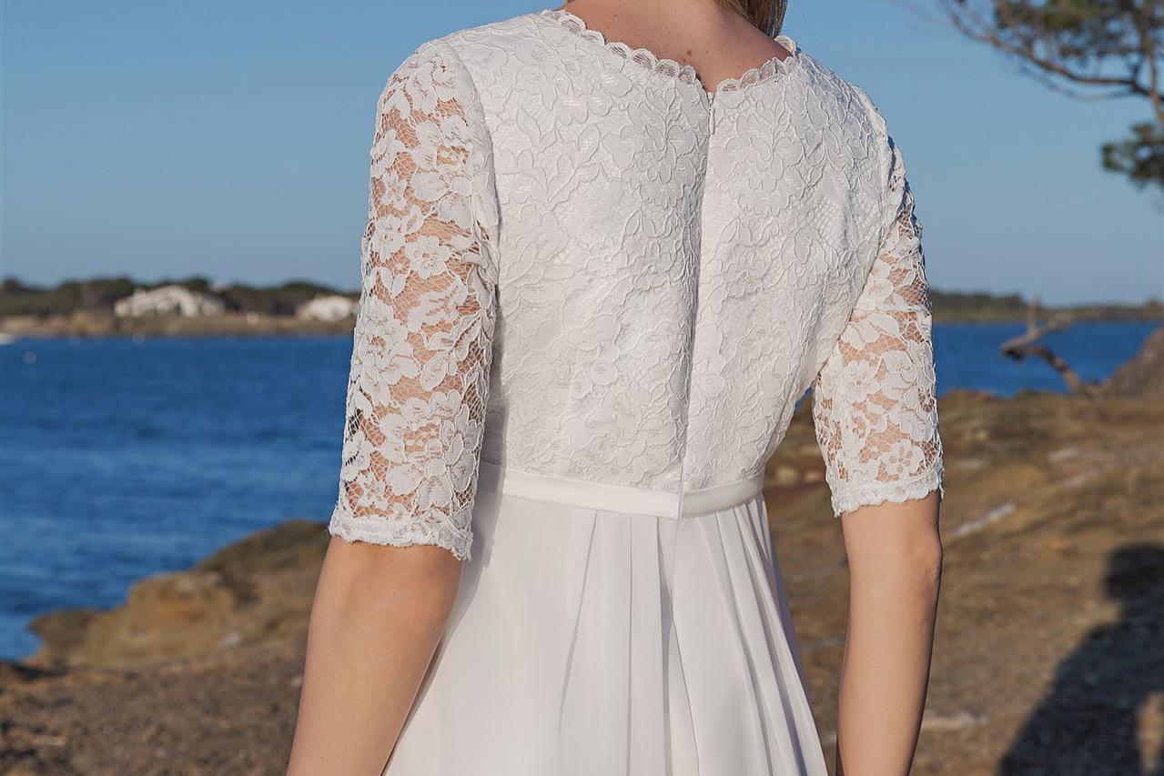 Eine blonde Frau in einem weißen Kleid steht an der felsigen Küste vor dem blauen Meer.