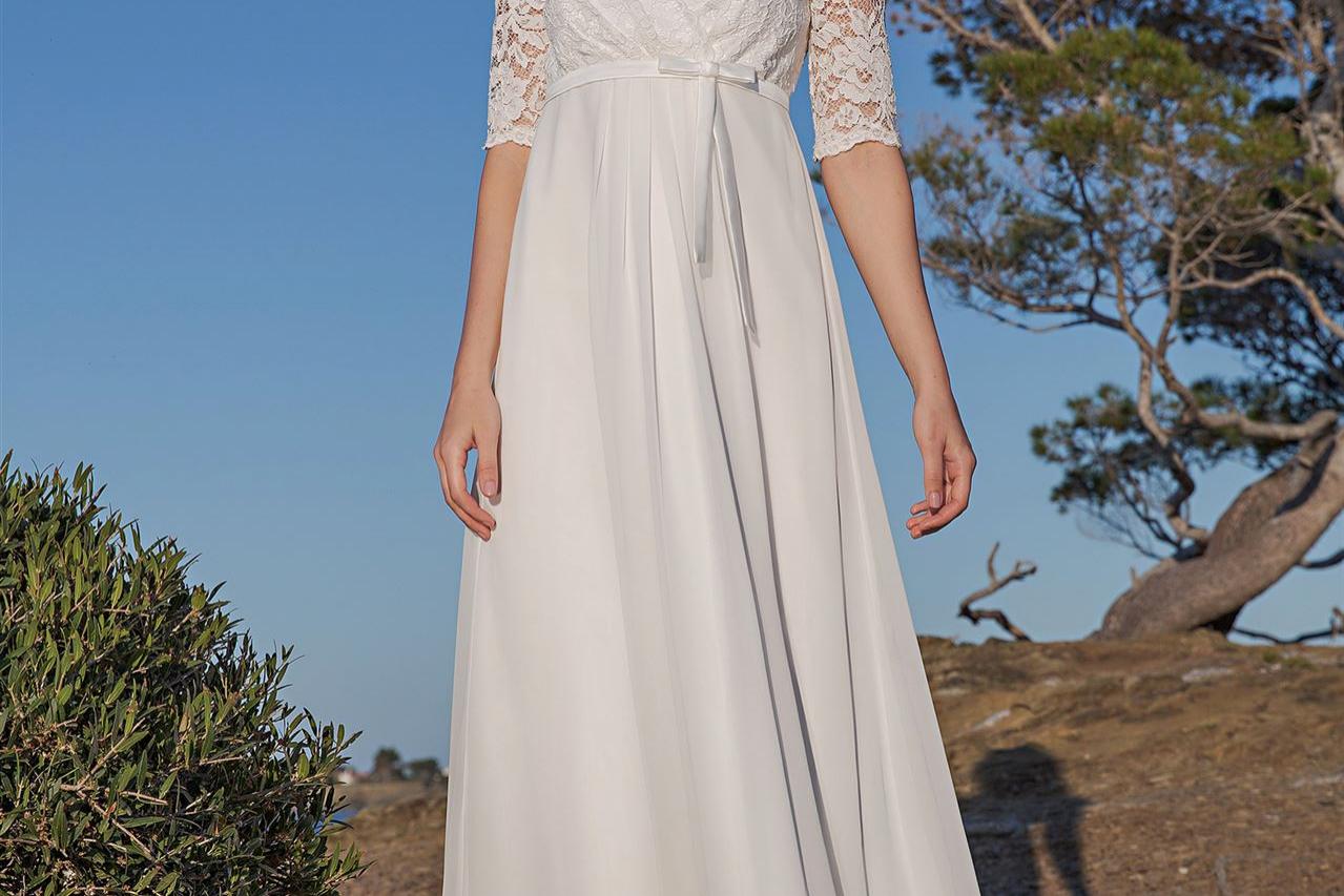 Blonde Frau in einem fließenden, hellen Sommerkleid steht draußen in einer natürlichen, sonnendurchfluteten Landschaft.