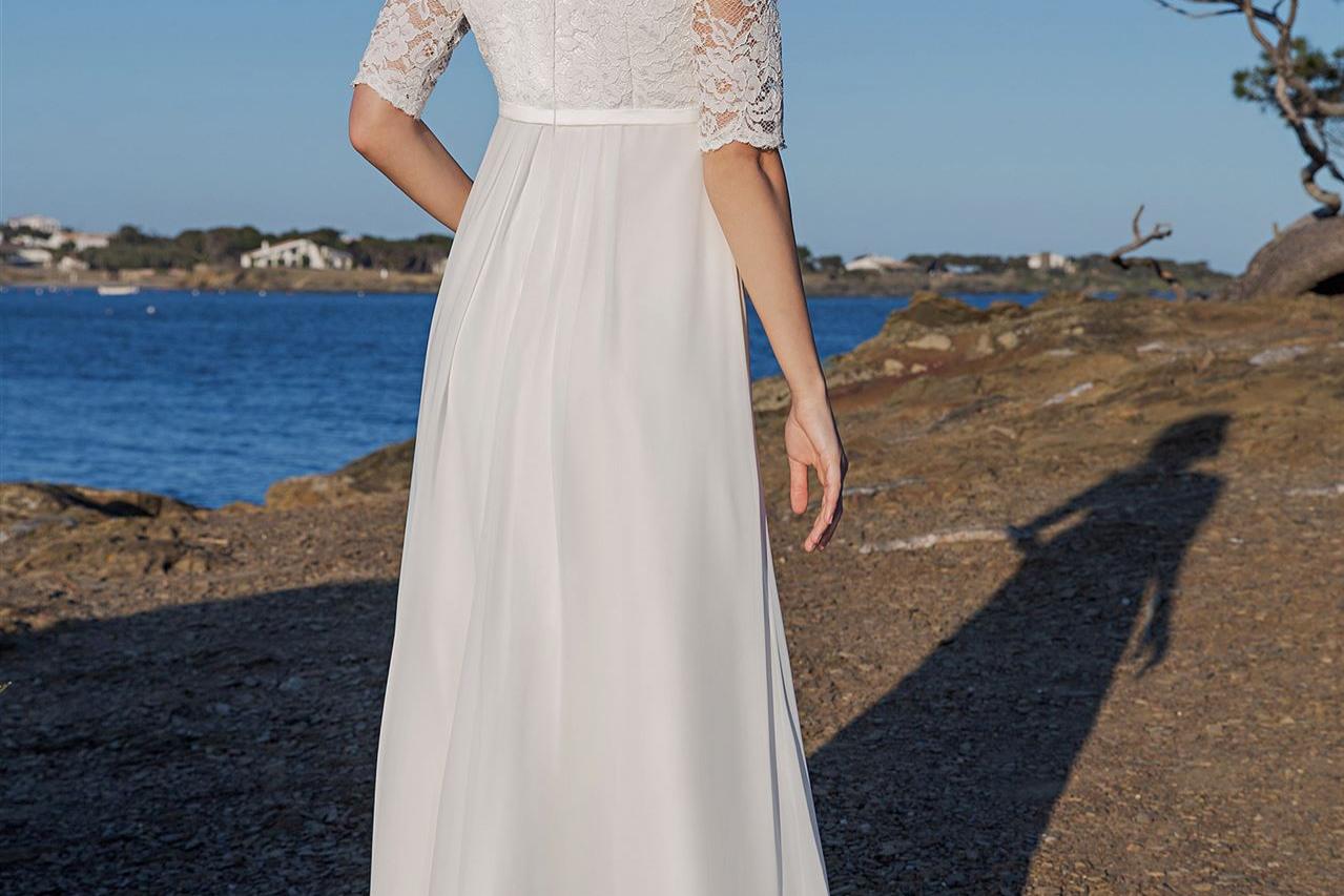 Blonde Frau in einem weißen, fließenden Kleid steht an einer steinigen Küste vor dem weiten Meer.