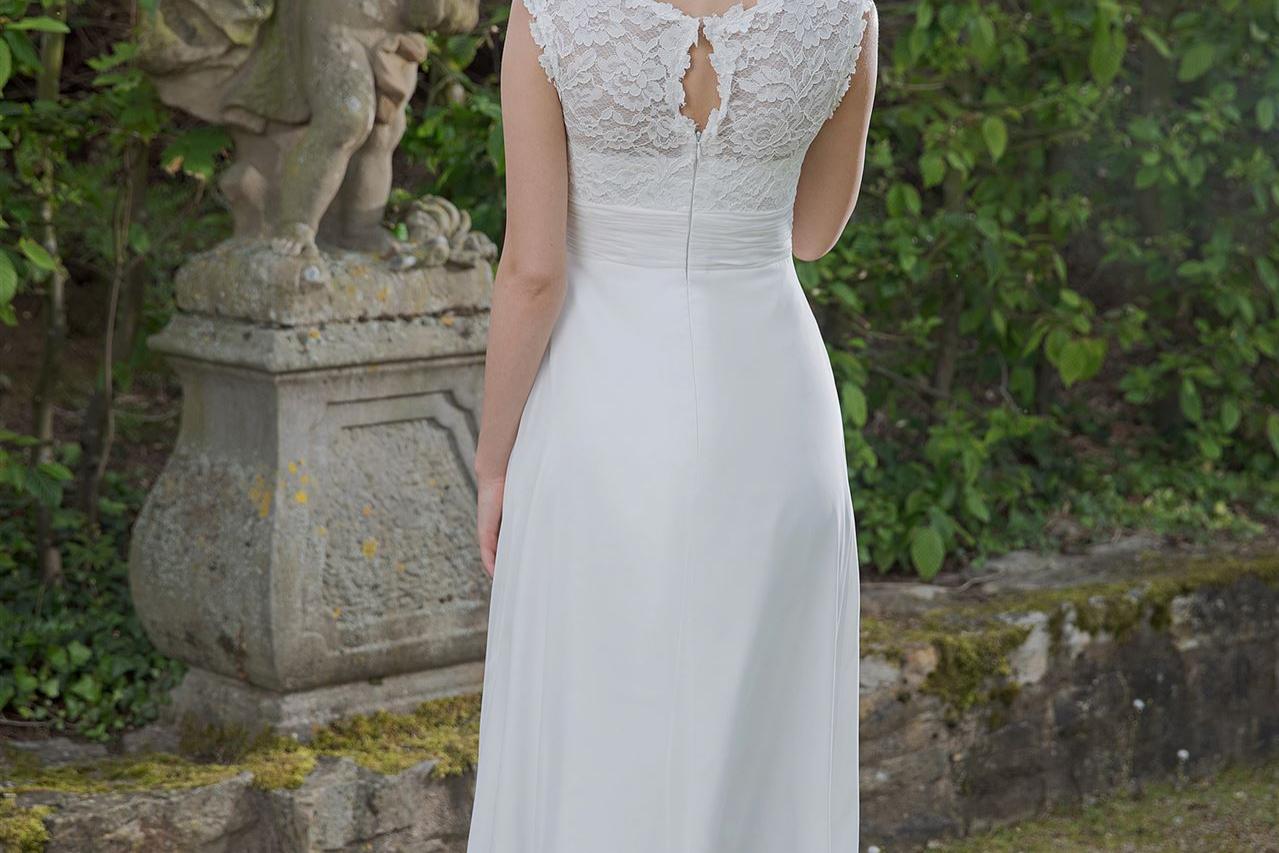 Eine blonde Frau in einem langen, weißen Kleid steht in einem Garten und wird von einer klassischen Steinstatue umgeben.
