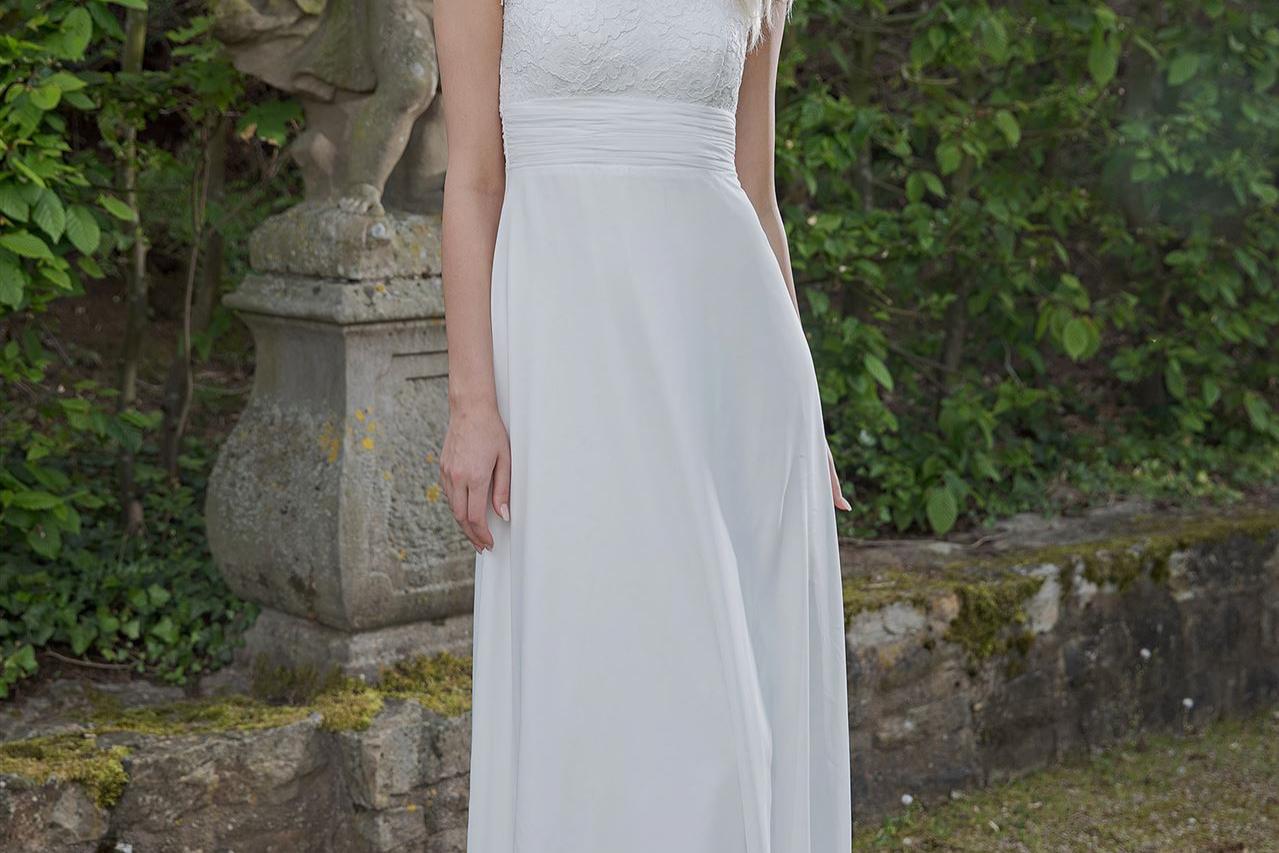 Blonde Frau in weißem Kleid vor einer steinernen Statue in einem grünen Garten.