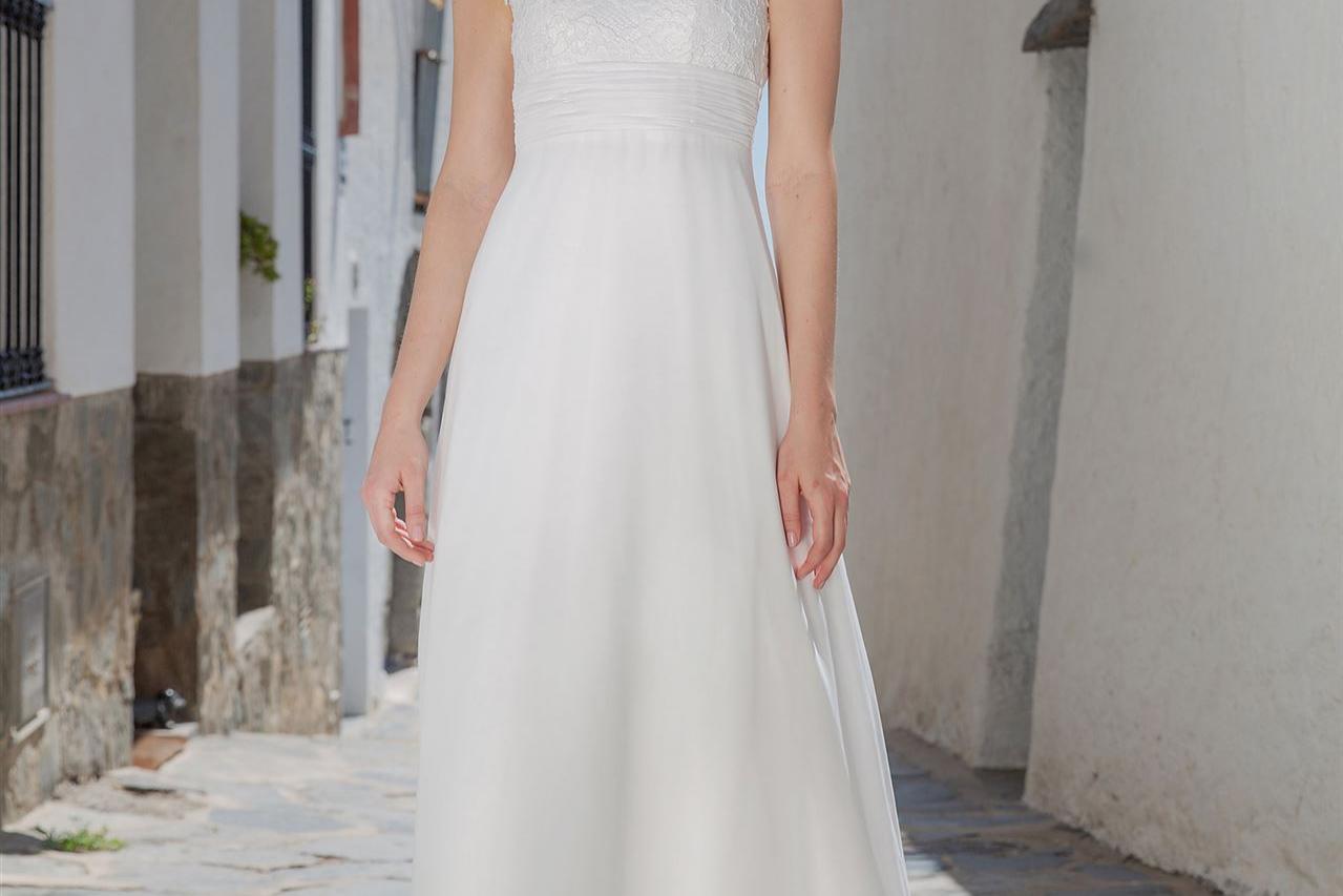 Eine junge Frau in einem eleganten, weißen Sommerkleid steht auf einem steinernen Weg zwischen historischen Gebäuden und einer weißen Mauer.