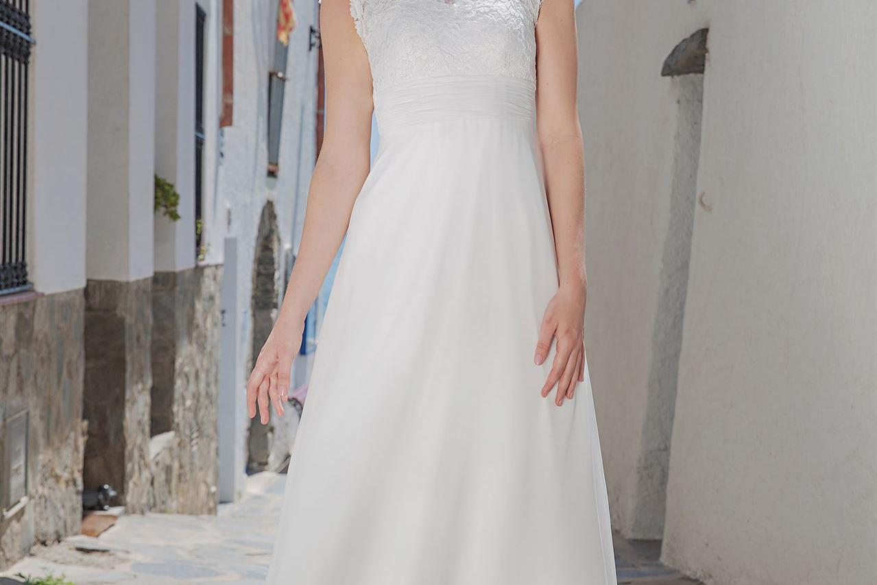 Eine blonde Frau in einem langen, weißen Kleid steht auf einem historischen Kopfsteinpflaster zwischen Gebäuden und einer weißen Wand.