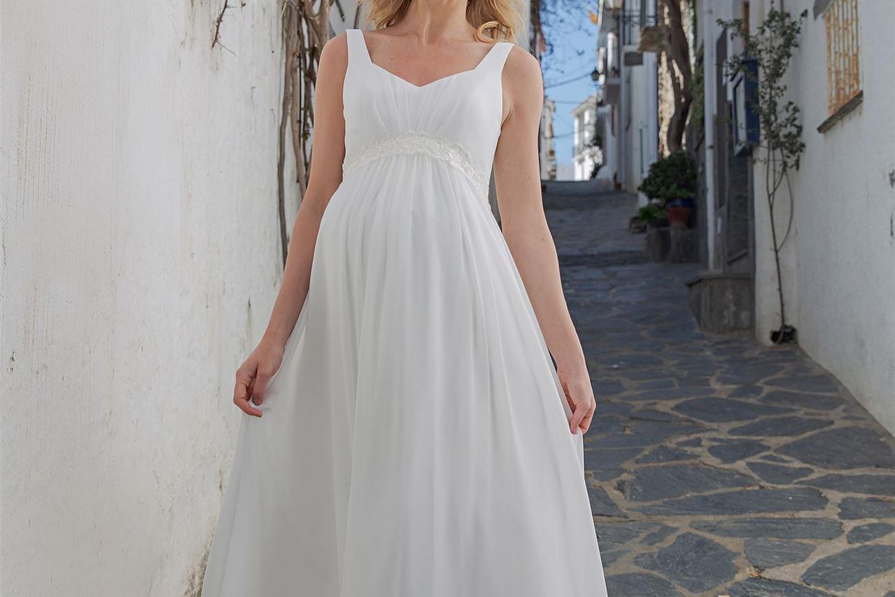 Junge Frau in einem fließenden weißen Kleid in einer sonnigen, mediterranen Gasse.