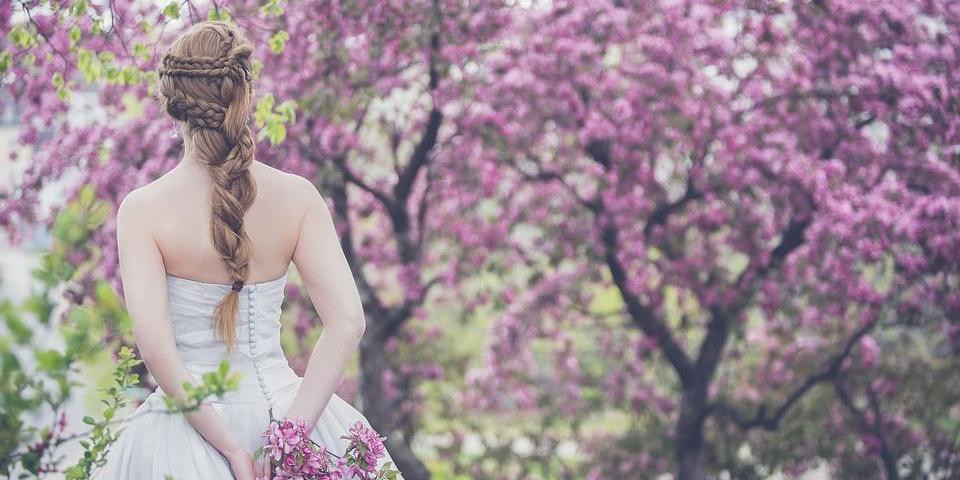 Braut in Vintage-Brautkleid und aufwändiger Flechtfrisur steht unter blühendem Apfelbaum. In der Hand hält sie einen Blütenzweig.